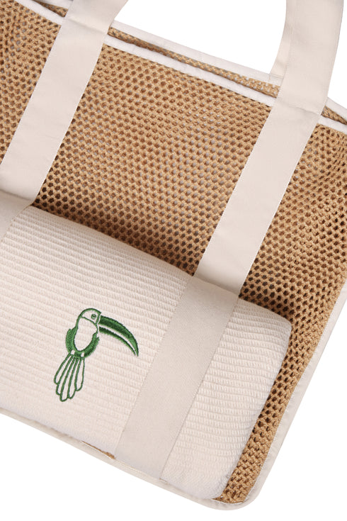 Close-up of a tan mesh beach bag with white straps holding a rolled white towel with a green toucan.